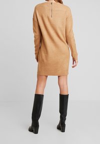 Robe tricotée couleur sable avec un col rond, des manches longues et une fermeture éclair au dos. Portée avec des bottes en cuir noires jusqu'aux genoux.