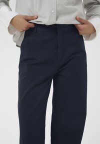 Mains ajustant la ceinture d'un pantalon bleu marine porté avec une chemise à manches longues de couleur claire sur un fond uni.