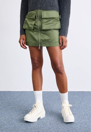 Gonna verde con zip e due tasche frontali, abbinata a calzini bianchi alla caviglia e anfibi bianchi chunky. Maglione grigio a trama visibile sopra.