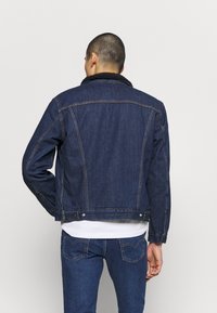 Denim jacket in dark blue with a sherpa-lined collar. Features visible stitching and metal hardware. Classic design with a tailored fit.