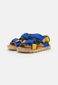 Blå og gule tekstil sandaler med justerbare stropper, teksturerte overflater, brun innersåle og en chunky beigefarget gummisåle.