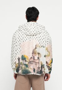 Hettegenser med hvitt stoff som har et logomønster. Baksiden viser en fargerik, kunstnerisk bysilhuett.