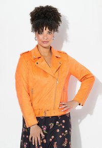 Veste de moto en simili cuir orange avec poches zippées, matériel argenté, silhouette ajustée et taille ceinturée, associée à une jupe à fleurs.