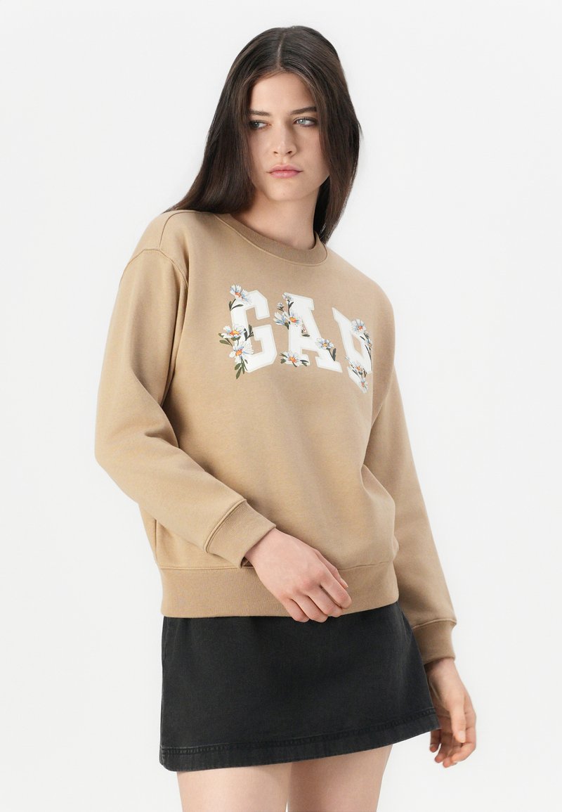 Joven mujer vestida con un sudadera beige de GAP con detalles florales y una falda negra, de pie contra un fondo blanco liso.