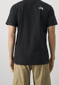 Czarny bawełniany t-shirt z krótkimi rękawkami, z małym białym logo na górnej części pleców. Noszony z beżowymi spodniami typu cargo.