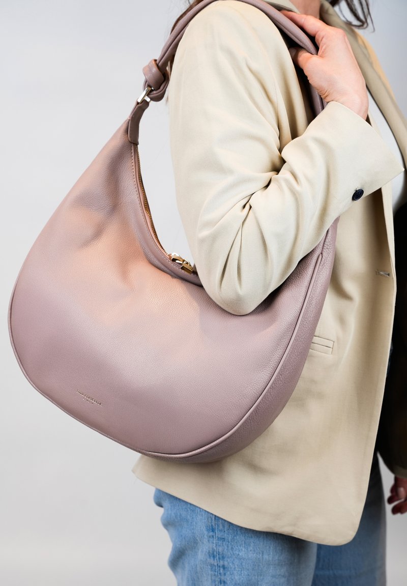 Lichtroze leren schoudertas met een gladde textuur, gebogen vorm en een afneembare strap, vastgehouden door een persoon in een beige jasje.