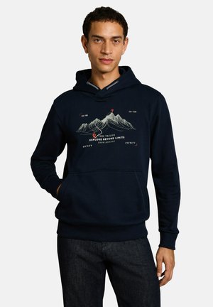 Junger Mann trägt einen dunkelblauen Kapuzenpullover mit Berggrafik und Text "Explore Beyond Limits" und dunkle Jeans vor einfarbigem Hintergrund.