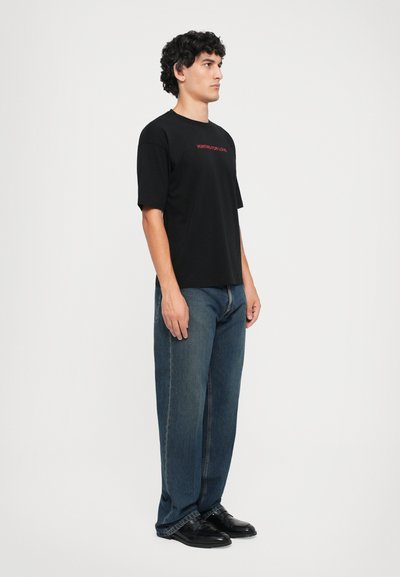 Svart bomull t-skjorte med korte ermer, med rød tekst "HUNTING FOR LOVE." Kombinert med løstsittende blå jeans og svarte sko.