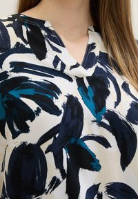 Blusa senza maniche in tessuto leggero, caratterizzata da audaci motivi a pennellate blu scuro e teal su uno sfondo crema, con scollatura a V.