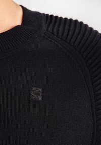 Mörk marinblå stickad tröja med ribbat mönster på axlarna och en liten präglad logotyp på bröstet, slät kropp med rundad halsringning.