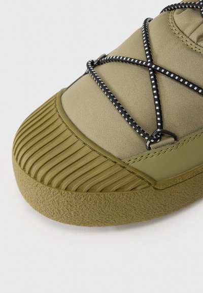 Olive roheline sneaker, millel on tekstuuriga kummitald, sile kangast ülemine osa ja väljapaistev must-valge bungee-köie sidumise süsteem.