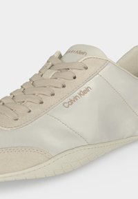 Μπεζ αθλητικό παπούτσι Calvin Klein με λεπτομέρειες από σουέτ και δέρμα, μπεζ κορδόνια και υφασμένο πάτο, εμφανίζεται από κοντινή πλευρική όψη.