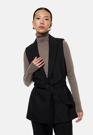 Kobieta nosząca złote kolczyki koła, golf taupe, czarną bezrękawnikową kamizelkę z paskiem, pozująca z ręką przy piersi na białym tle.