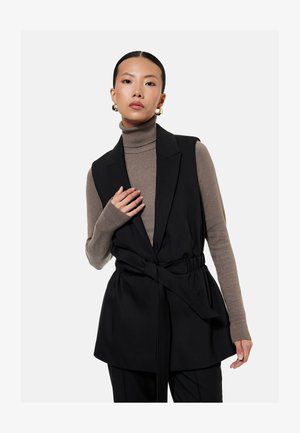 Kvinde iført guldbøjleøreringe, taupe rullekravesweater, sort ærmeløs vest med bælte, poserer med hånd nær brystet mod hvid baggrund.