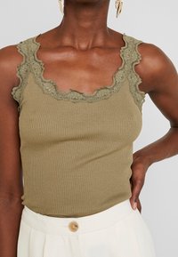 Dark-skinned person wearing olive green ribbed tank top with lace trim and white high-waisted pants with button, hand on hip.