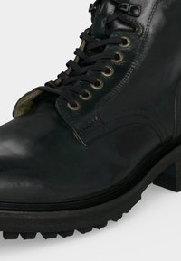 Botte noire en cuir à lacets avec œillets métalliques et semelle épaisse en caoutchouc, montrée en gros plan avec un focus sur les détails avant et latéraux.