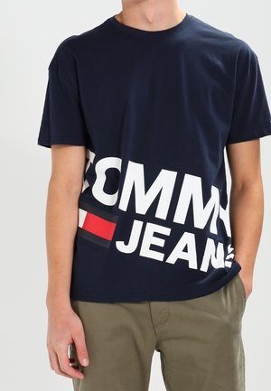 Osoba ubrana w granatowy t-shirt Tommy Jeans z dużym białym i czerwonym logo, w połączeniu z oliwkowozielonymi spodniami.