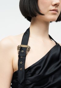 Κοντινό πλάνο ενός ατόμου που φοράει μαύρο σατέν τοπ με λουρί που καταλήγει σε διακοσμητική χρυσή αγκράφα με την επιγραφή "Versace Jeans Couture".