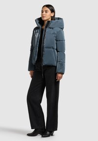 Paminkštinta, su gaubtu striukė akmens mėlynos spalvos, su užtrauktuku, blizgiomis detalėmis ir laisvo silueto. Dėvima su tamsiomis kelnėmis ir batais.