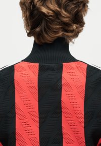 Tagakülje pealt vaadatav inimene laineliste pruunide juustega, kandes musta ja punase triibulise mustriga kampsunit, millel on tekstureeritud mustrid ja kõrge soonikuga krae.