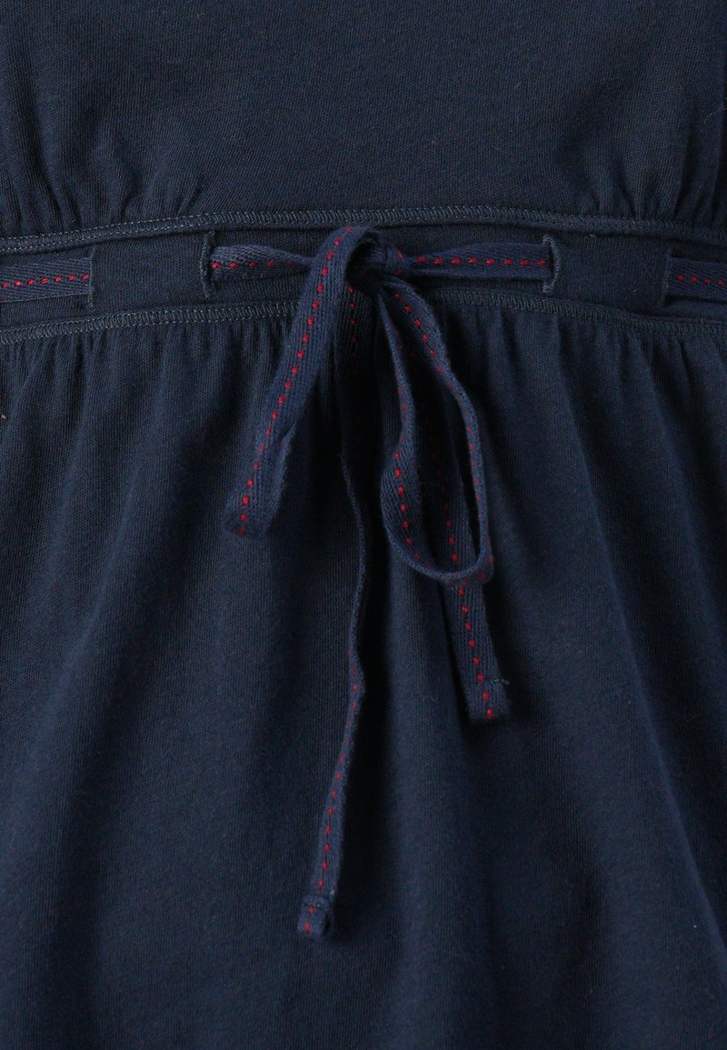 Vestido azul marino con cintura fruncida, que presenta un lazo cosido en rojo y una cinturilla elástica. Fabricado en un suave material de algodón.