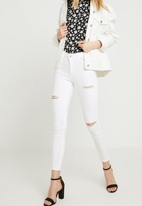 Jeans strappati bianchi con una vestibilità slim, abbinati a un top floreale nero e una giacca bomber bianca, completati con tacchi con cinturino alla caviglia neri.