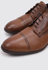 Zapatos de cuero marrón con punta redondeada, acabado de bajo brillo y diseño clásico de cordones. Presentan detalles de costura y una suela negra.