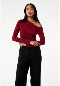 Bordeaux langærmet top med off-shoulder design og gathered tekstur, parret med højhentede sorte bukser.