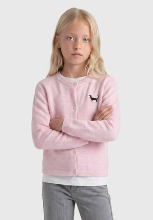 Jeune fille aux longs cheveux blonds, portant un cardigan rose avec un emblème de chien noir, chemise blanche et pantalon gris, bras croisés, expression neutre.