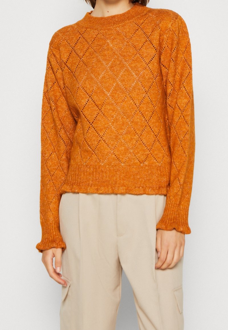 Femme portant un pull orange tricoté avec un motif en losange et un pantalon beige sur un fond blanc uni.