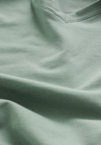 T-shirt vert clair en V en coton doux, présentant une texture lisse, des coutures discrètes et un tissu drapé montrant sa coupe décontractée.