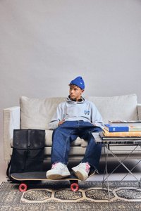 Un adolescente vestito con abiti casual e un berretto blu è seduto su un divano con un monopattino sotto i piedi, uno zaino nero nelle vicinanze e libri su un tavolino.