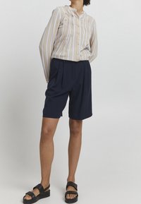 Camisa de color beige claro con rayas verticales azules, cuello abotonado y pliegues; combinada con pantalones cortos azul marino ajustados y sandalias negras.