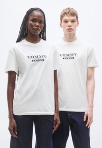 Λευκά βαμβακερά μπλουζάκια με το λογότυπο "TOMMY JEANS" σε μαύρο, στρογγυλή λαιμόκοψη, κοντά μανίκια, με σιλουέτες μοντέλων σε απλό φόντο.