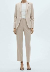 Blazer beige sur mesure et pantalon assorti, en tissu lisse. Le blazer est doté d'un bouton unique et d'une poche. Associé à un haut blanc.