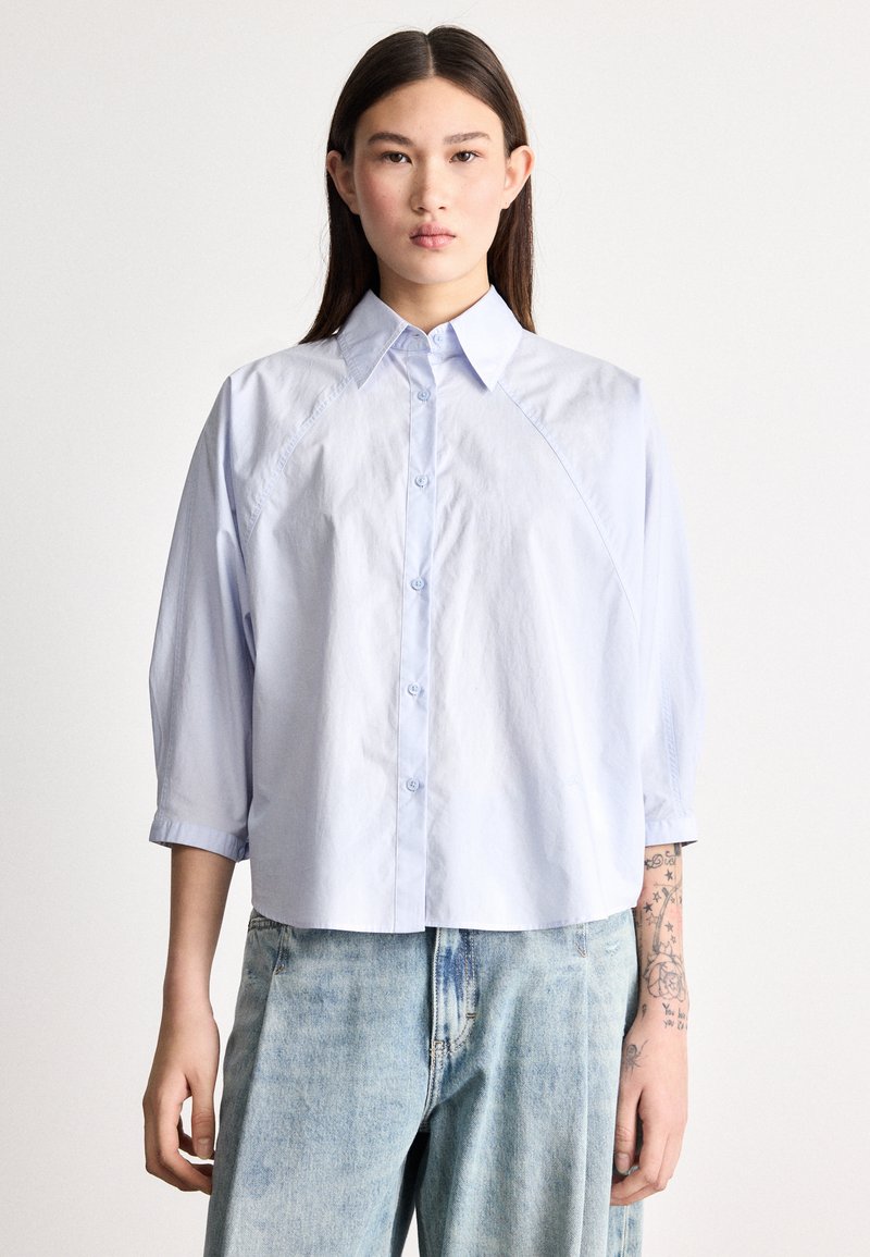 Ανοιχτό μπλε, ελαφρύ πουκάμισο με κουμπιά μπροστά, γιακά και μανίκια τριών τετάρτων. Ομαλή υφή και άνετη εφαρμογή.