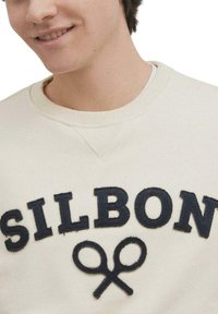 Sudadera de color crema claro con texto bordado en navy "SILBON" y un motivo de tijeras. Presenta un cuello redondo y puños de canalé. Textura suave.