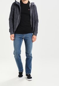 Felpa con zip di colore navy con rivestimento in pile bianco, t-shirt nera sotto, jeans blu e scarpe nere con suola bianca, in piedi su sfondo bianco.