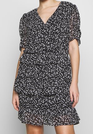 Femme portant une robe noire à imprimé floral blanc, col en V, manches courtes bouffantes et jupe à volants superposés.