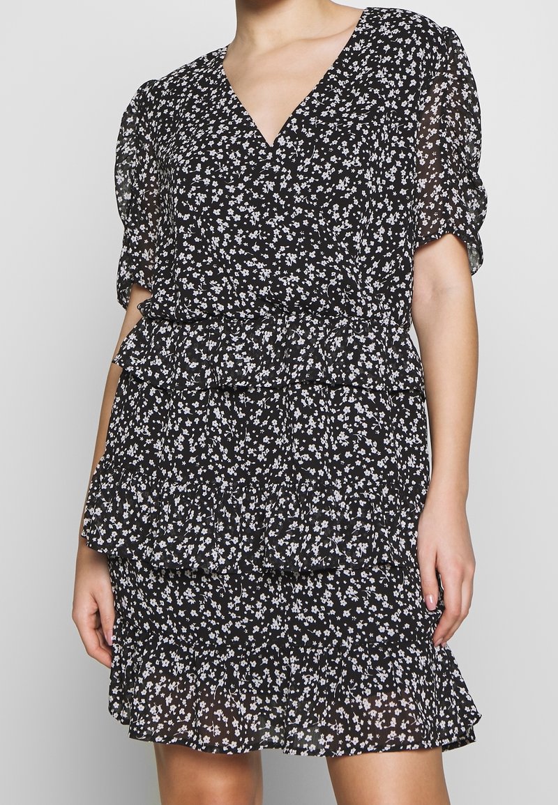 Femme portant une robe noire à imprimé floral blanc, col en V, manches courtes bouffantes et jupe à volants superposés.