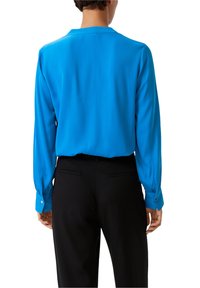 Camicia a maniche lunghe di un blu brillante, con una texture liscia e un colletto classico. Presenta polsini con bottone e una vestibilità rilassata. Indossata con pantaloni neri.