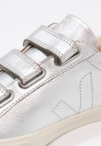 Baskets en cuir argenté avec deux sangles velcro, présentant une finition texturée, un bout rond et une semelle en caoutchouc avec des détails de design minimalistes.