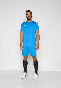 Lyseblå sports skjorte og shorts med sorte knæhøje sokker og hvide sportssko. Har Under Armour-logoet i gul på brystet og shorts.