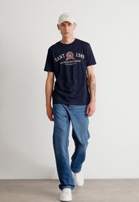 Tricou navy cu grafică "GANT 1949", blugi albaștri, adidași albi și o șapcă de culoare deschisă; croi lejer cu mâneci scurte și picior drept.
