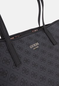 Sac cabas noir Guess avec motif de logo en relief, anses en cuir et inscriptions dorées "GUESS EST 1981" sur l'extérieur en tissu.