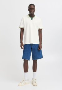 Camiseta tipo polo en color crema con cuello verde y puños a rayas, combinada con pantalones cortos de mezclilla azules y zapatillas blancas, con tejido texturizado.