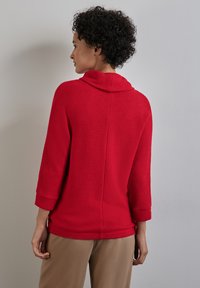Maglione rosso lavorato a maglia con colletto alto, maniche a tre quarti e un motivo testurizzato. Un dettaglio di cucitura corre verticalmente lungo la schiena.