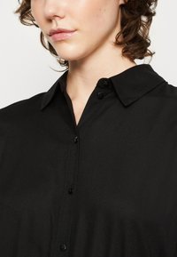 Camicia nera con colletto, caratterizzata da una texture liscia con bottoni neri, design ben modellato e un leggero lucore.