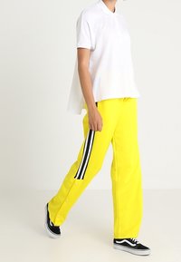 Camisa polo blanca combinada con pantalones anchos amarillos brillantes que cuentan con rayas laterales negras y detalles de botones a presión. Zapatillas negras completan el conjunto.