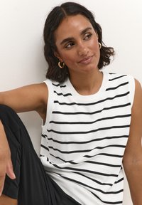 Haut sans manches blanc avec des rayures horizontales noires, col côtelé et texture lisse. Le modèle porte un pantalon noir et des boucles d'oreilles en or.